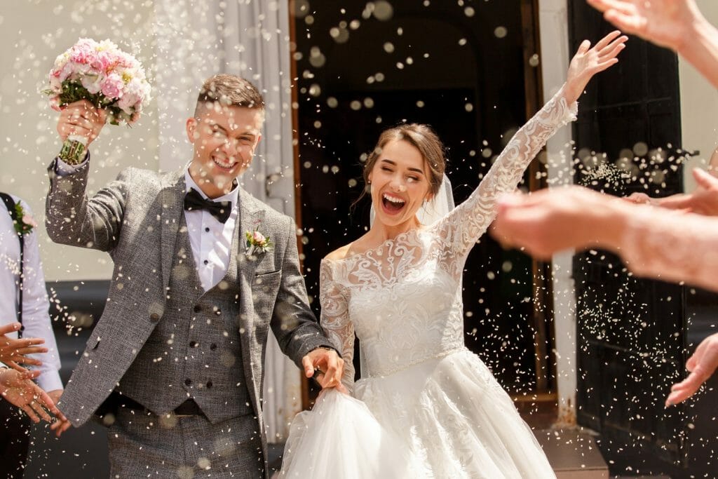 Une photo de mariés qui sortent de la mairie en souriant. Le marié tient un bouquet de fleurs dans la main droite.